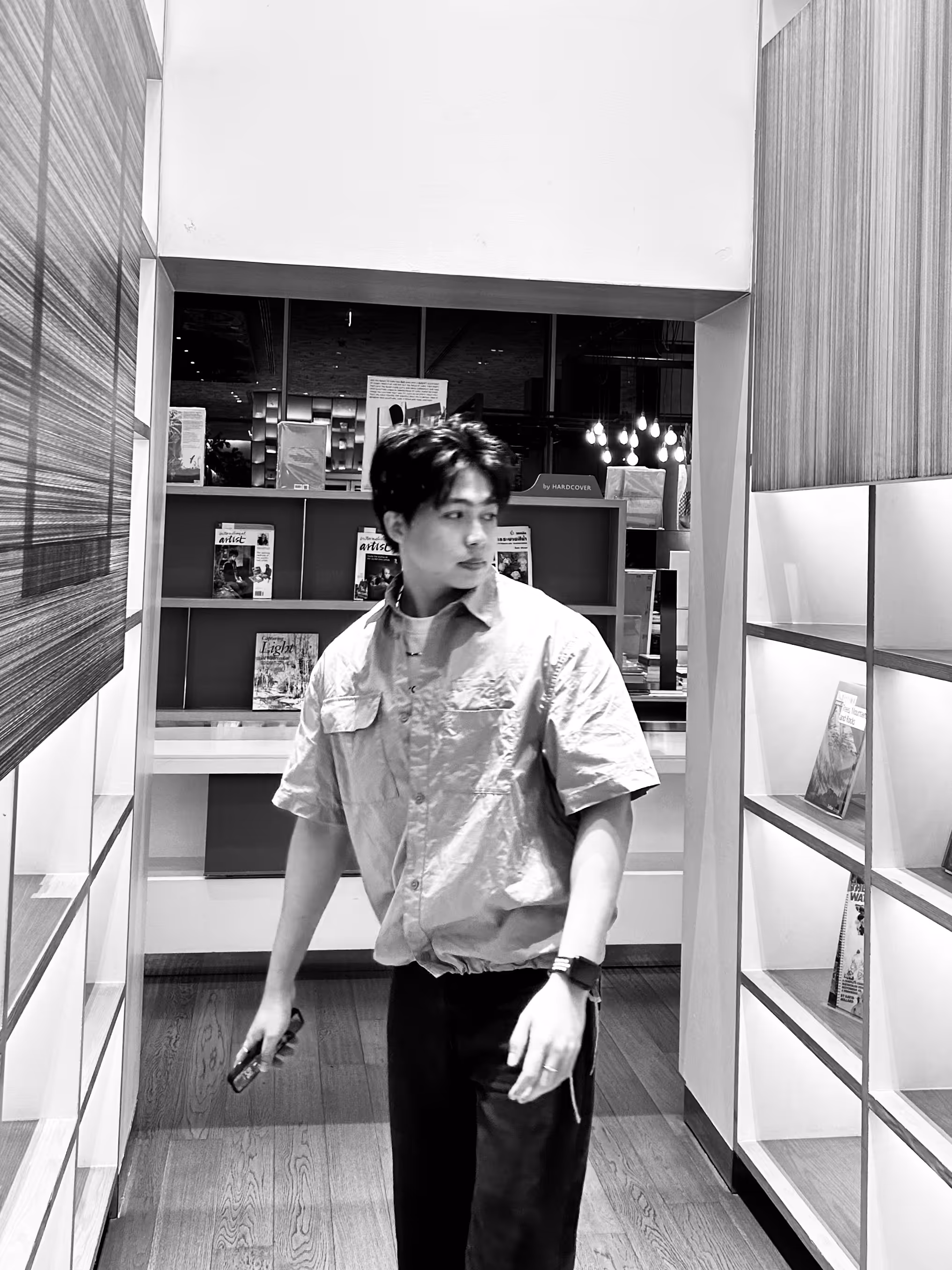 Young man in a short-sleeve shirt holding a smartphone, standing in a modern room with bookshelves.