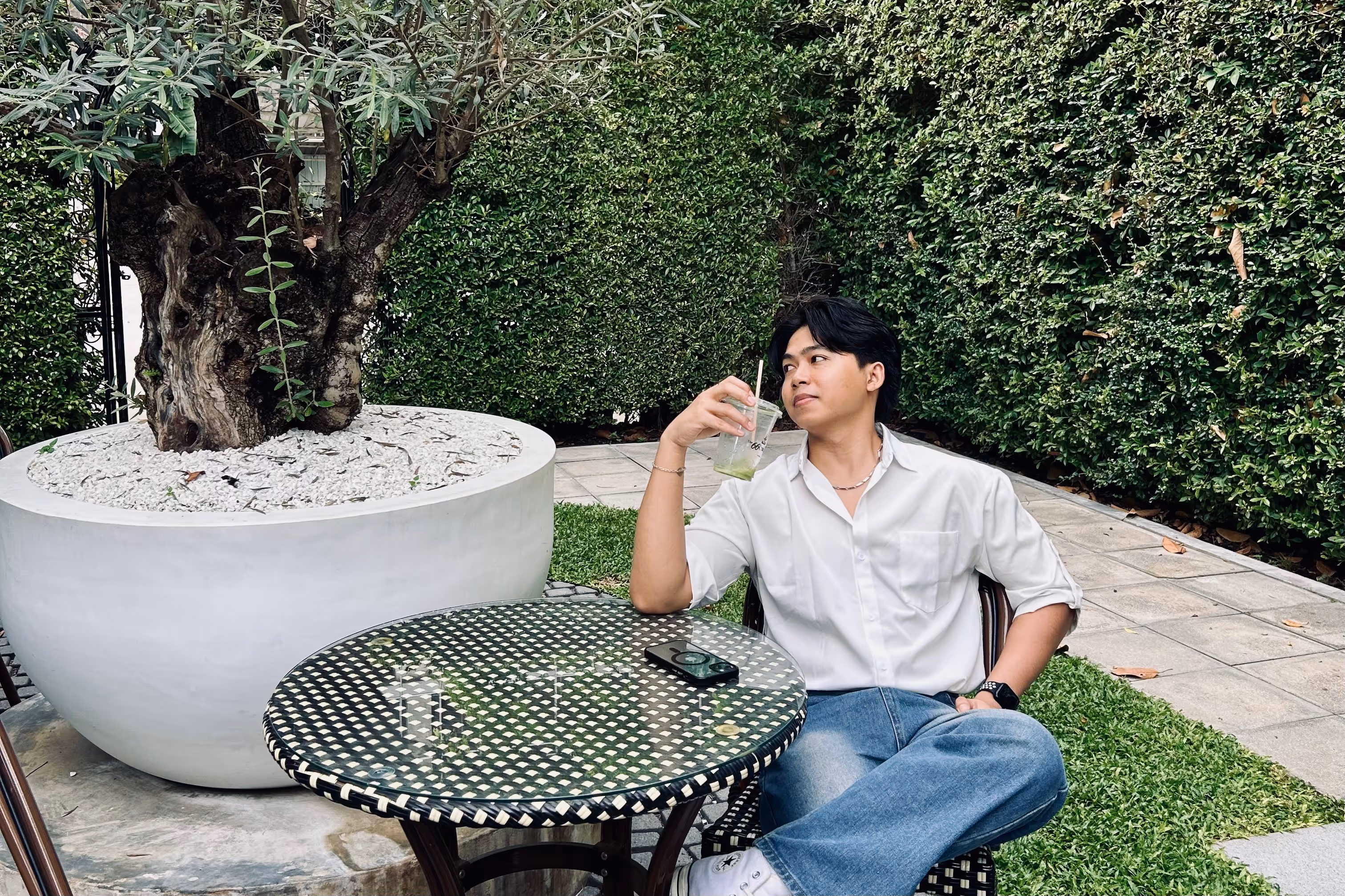 Young man in a white shirt and jeans sitting outdoors at a round wicker table, holding a drink and looking to the side.