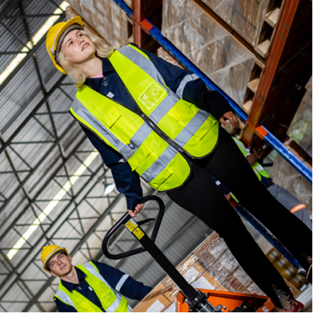 Zwei Lagerarbeiter mit gelben Schutzhelmen und reflektierenden Warnwesten arbeiten in einem Lager mit Regalen und Kartons.