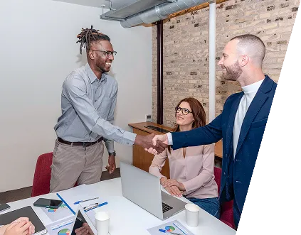 Zwei Männer schütteln sich lächelnd die Hand vor einer Frau, die am Tisch mit Laptop und Unterlagen sitzt.