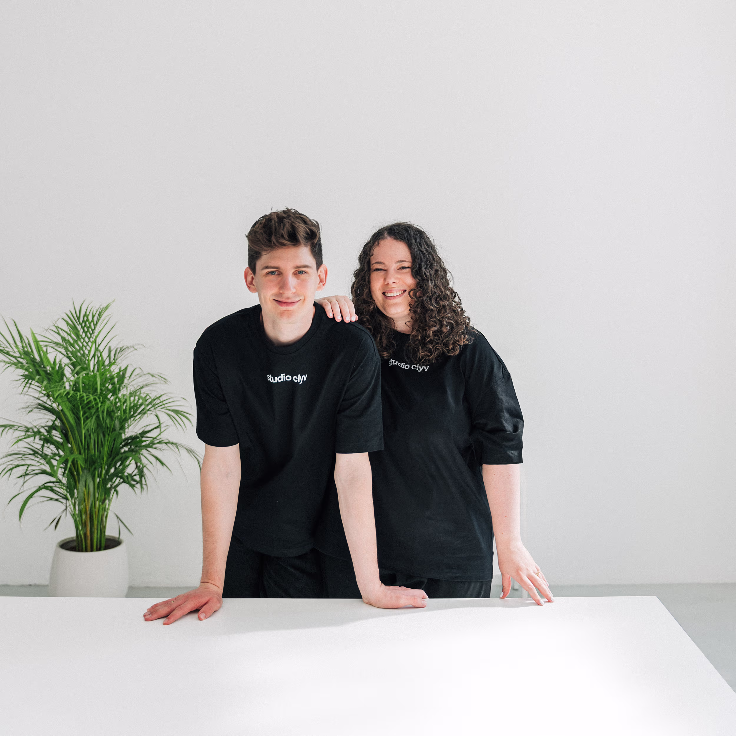 Zwei lächelnde Personen in schwarzen Studio-Clyv-T-Shirts lehnen auf einem weißen Tisch vor weißer Wand mit Topfpflanze links.