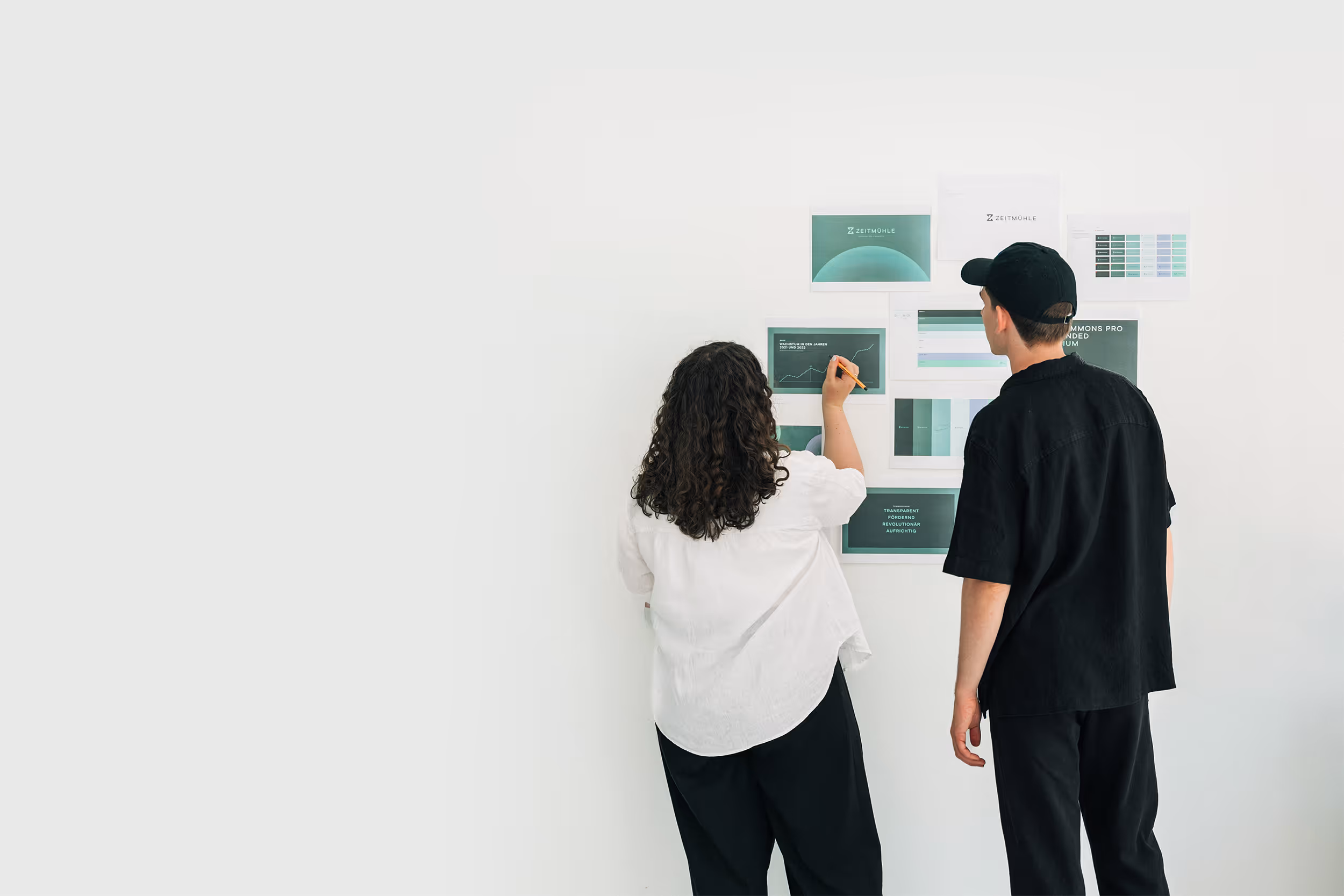 Eine Frau mit weißen Hemd und ein Mann mit schwarzem Hemd stehen vor einer Wand mit Grafiken und Diagrammen, die analysiert werden.