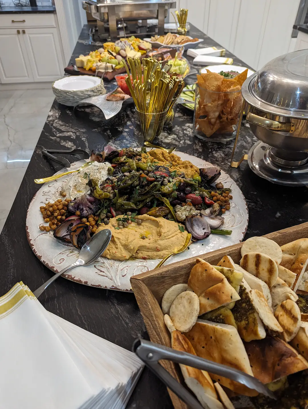 An artfully arranged table full of grazing board type appetizers. Starting closest, with a wooden bin with a wide variety of breads, next to a large white platter full of hummus, whipped feta, and babaganoush, each being cordoned off with a heap of grilled