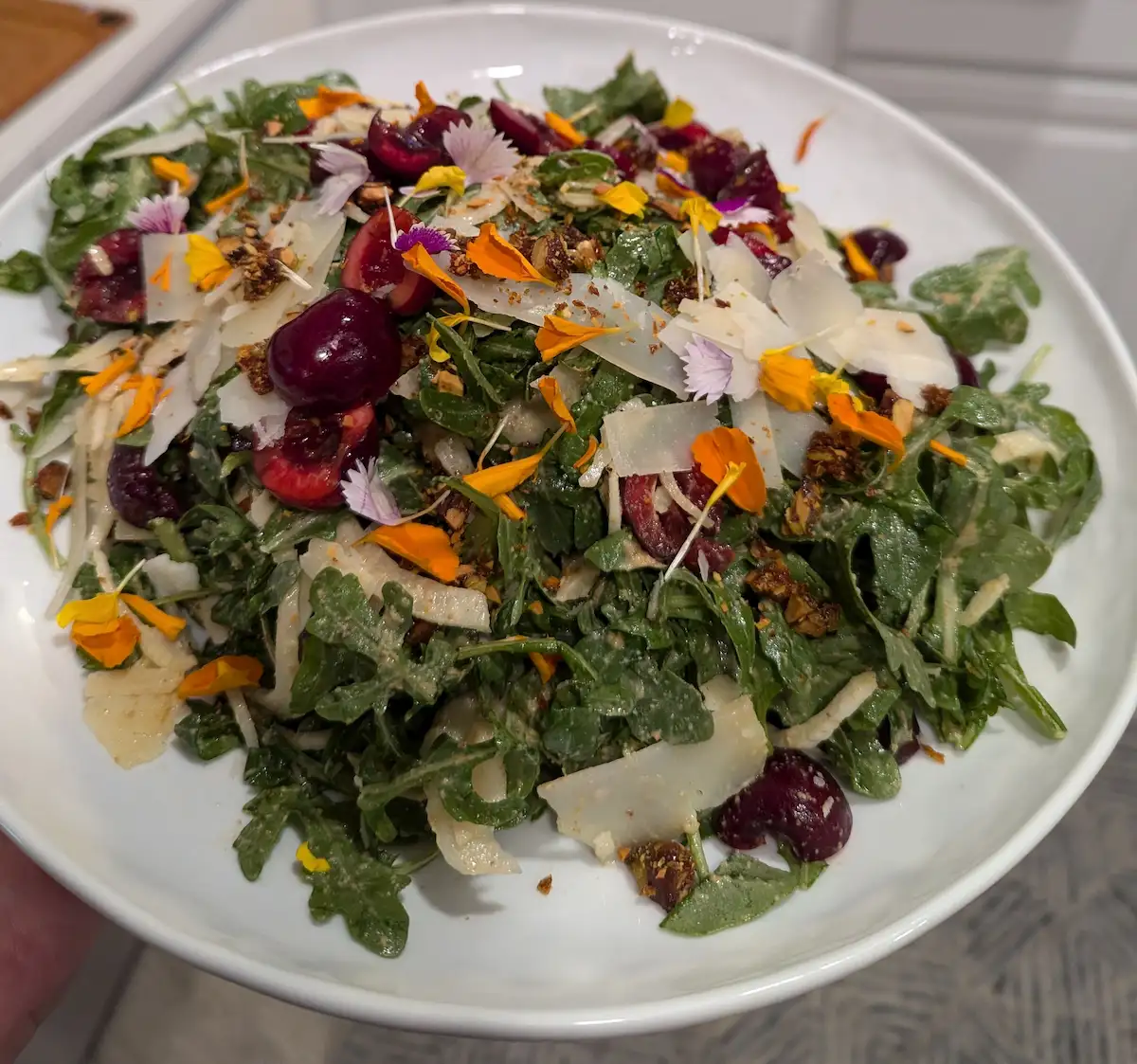 A plate full of beautifully dressed arugula that's been tossed with halved cherries, candied pistachios, shaved fennel, and shaved parmesan, topped off with a bright mix of edible flower petals