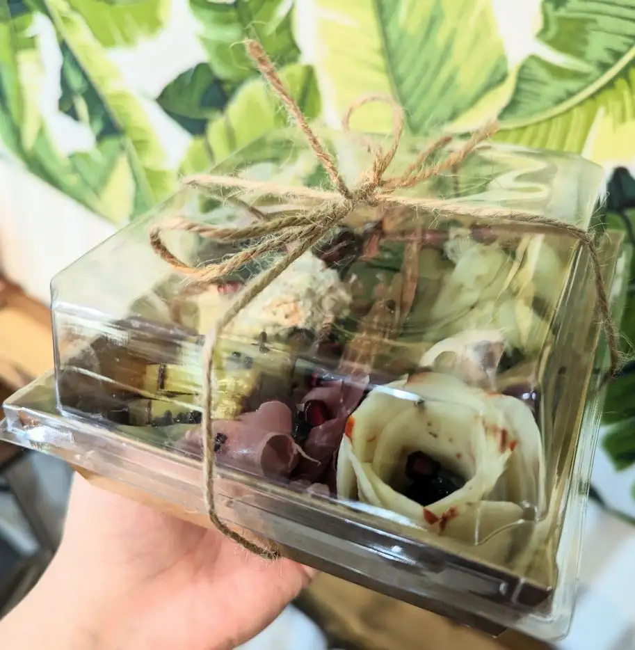A bamboo board covered with a variety of cured meats and cheeses, sprinkled through with grapes, cherries, and cantaloupe balls. Two small cups of olives are also included.