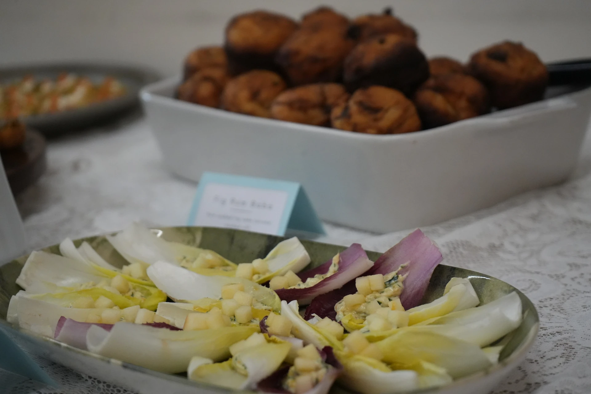 A plate of endive leaves, alternating purple and yellow, stuffed with finely diced apples and goat cheese. Behind this plate a white baking dish of muffins can be seen.