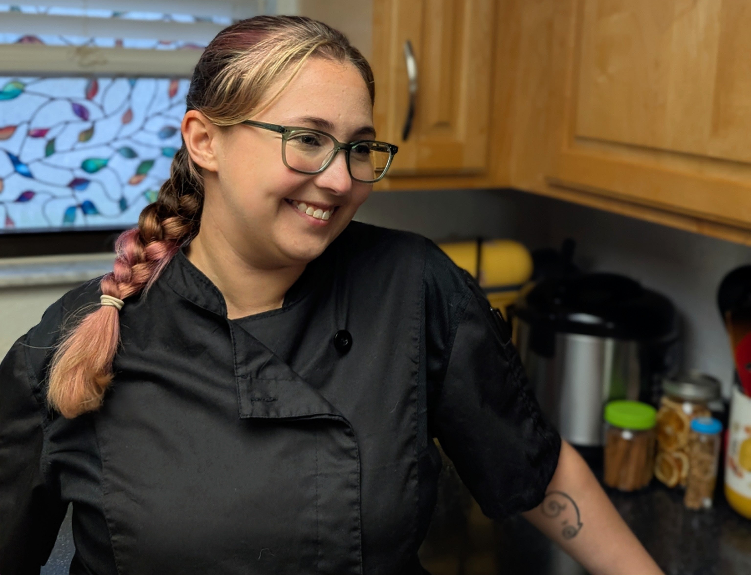 A female chef in a black uniform in front of jars of spices and cooking equipment.