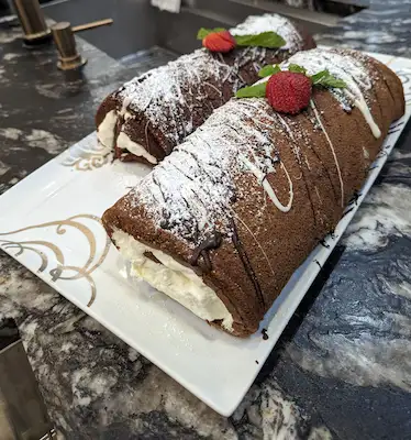 Two chocolate cakes rolled and filled with a whipped cream mixture, decorated with dark and white chocolate drizzles, fresh mint, and strawberries