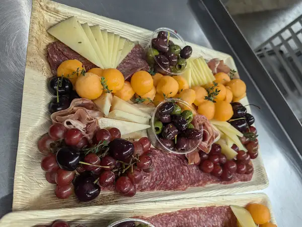 A bamboo board covered with a variety of cured meats and cheeses, sprinkled through with grapes, cherries, and cantaloupe balls. Two small cups of olives are also nestled in
