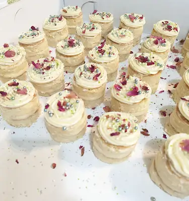 Twenty-five, one inch diameter vanilla cakes, made up of two tiers, topped with champagne buttercream rosettes, and sprinkled with crushed dried rose petals, and edible sugar pearls