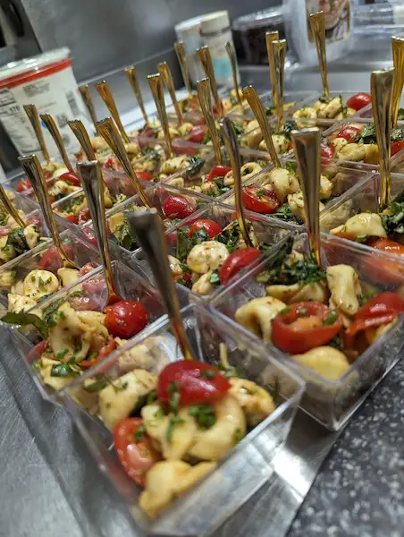Many clear square cups lined up, filled with halved cherry tomatoes and mozzarella pearls, tossed with a balsamic vinegar and lots of fresh herbs