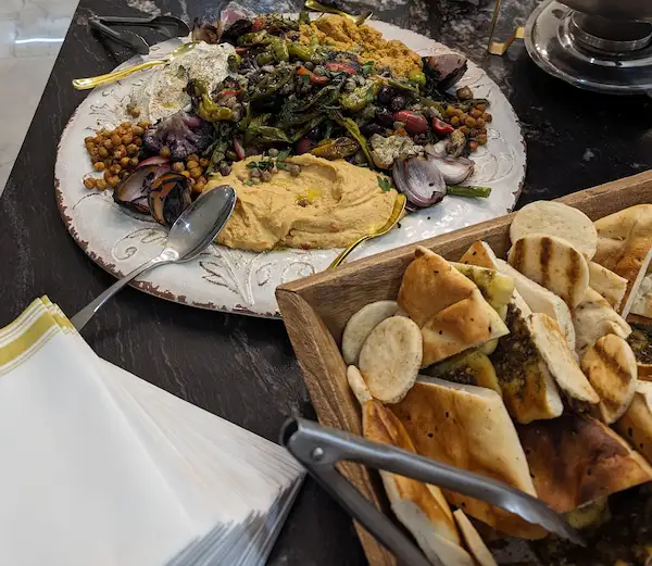 A wooden bin with a wide variety of breads, next to a large white platter full of hummus, whipped feta, and babaganoush, each being cordoned off with a heap of grilled vegetables and roasted chickpeas