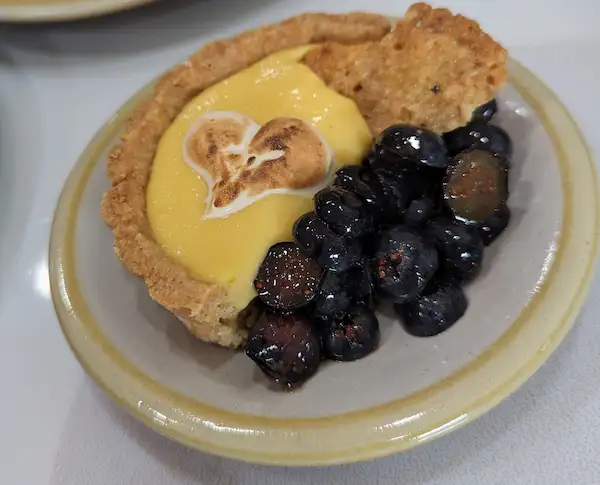 Half of a miniature lemon tart topped with a small meringue heart, that has been lightly toasted, with a side of macerated halved blueberries