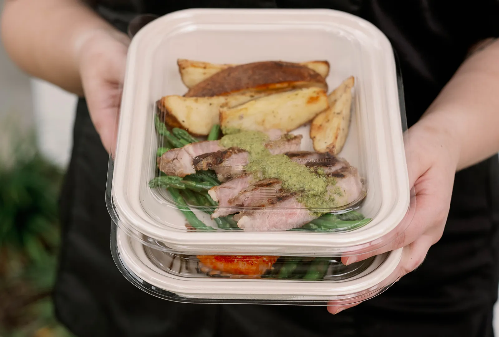 A chef holds two compostable boxes with clear lids. In the top box is sliced steak with chimichurri sauce, green beans, and roasted wedge potatoes.