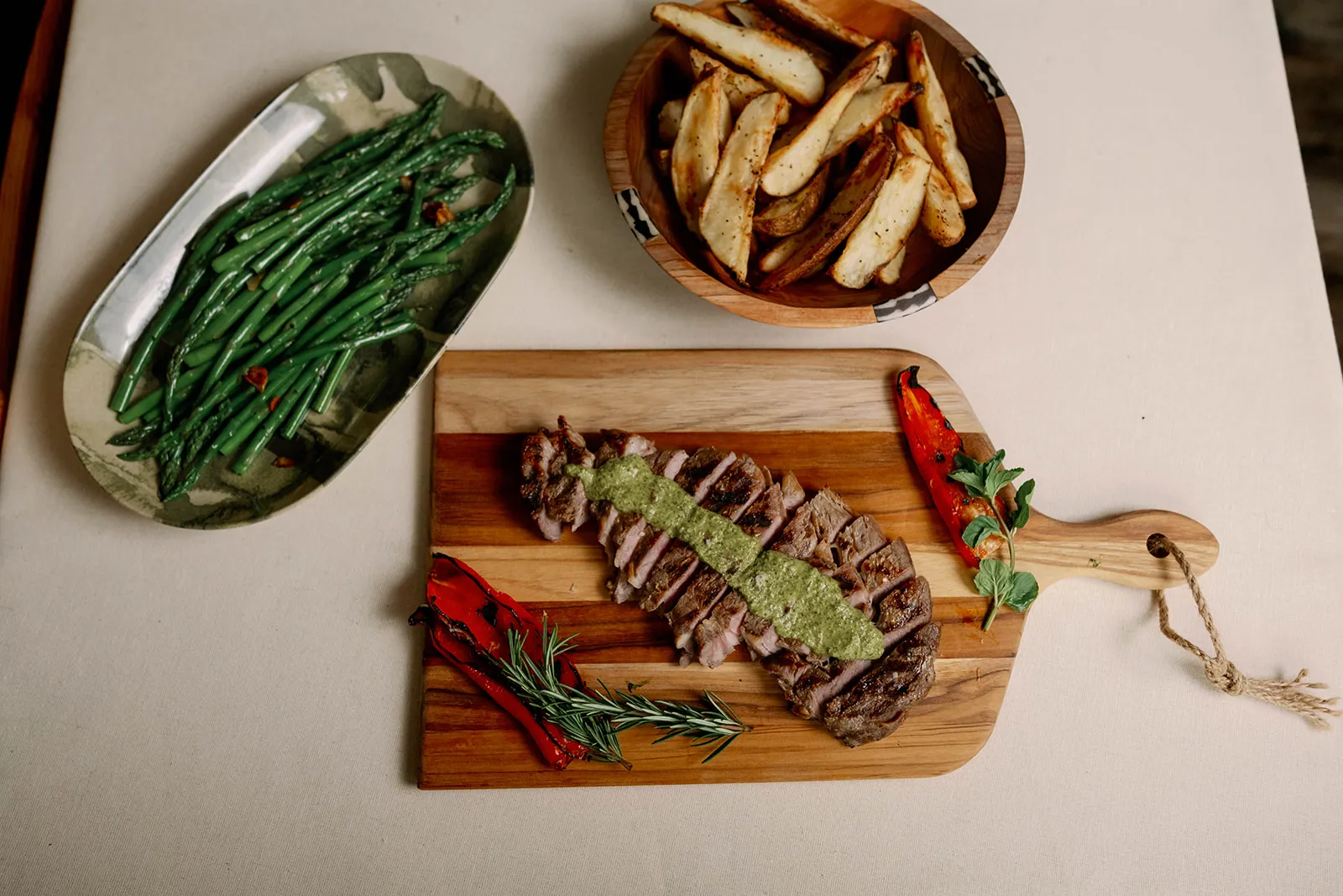 Top left- sauteed aspargus dotted through with toasted garlic, next to a wooden bowl of golden brown potato wedges, and a wooden booard featuring a grilled, sliced NY Strip steak topped with a chimichurri sauce