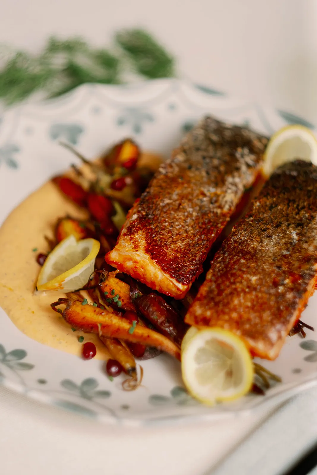 Red harissa salmon featuring crispy pan seared skin, yellow lemons, roasted carrots and a red pepper labneh sit on a delicate white plate.