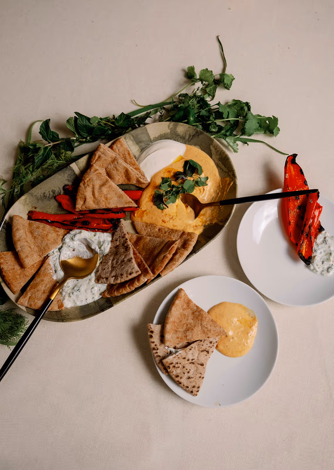 A serving platter and plate with whipped fetas, tzatziki, warm pita, and roasted peppers