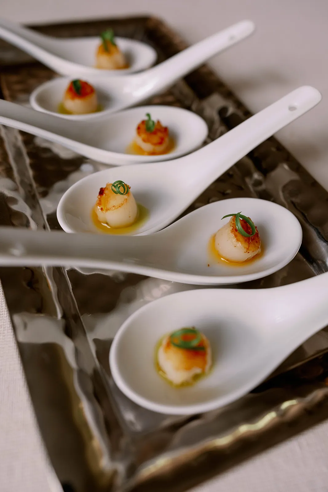 Seared scallops garnished with green basil oil rest in large white spoons on a serving platter