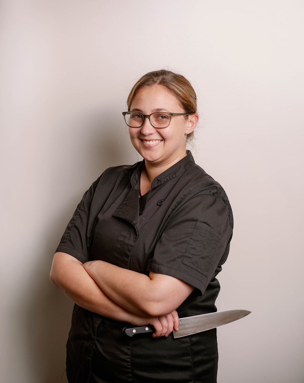 A smiling female chef in a black uniform stands in front of a white backdrop holding a chef’s knife.
