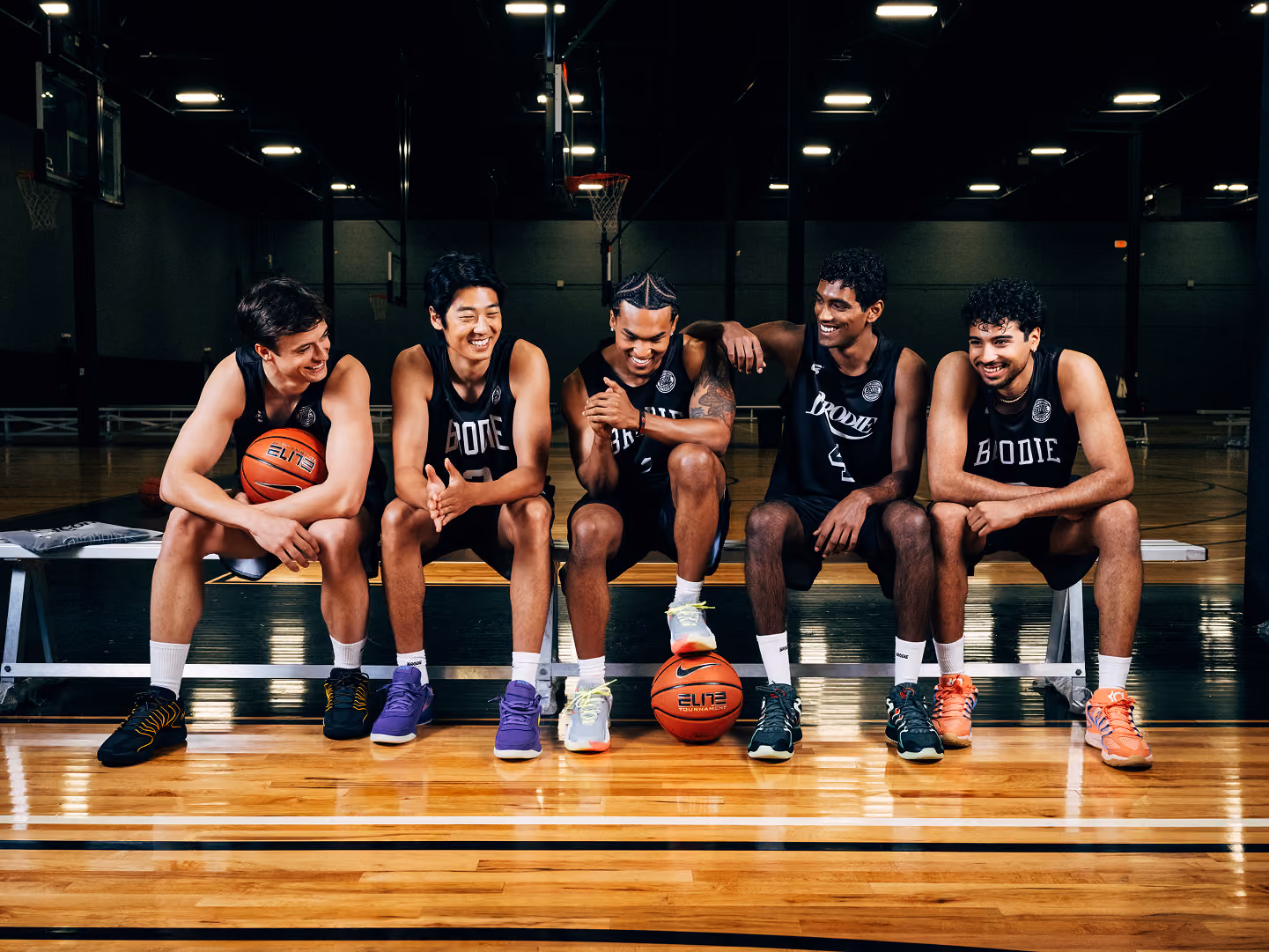 Five basketball players in black Brodie jerseys holding basketballs on an indoor court.