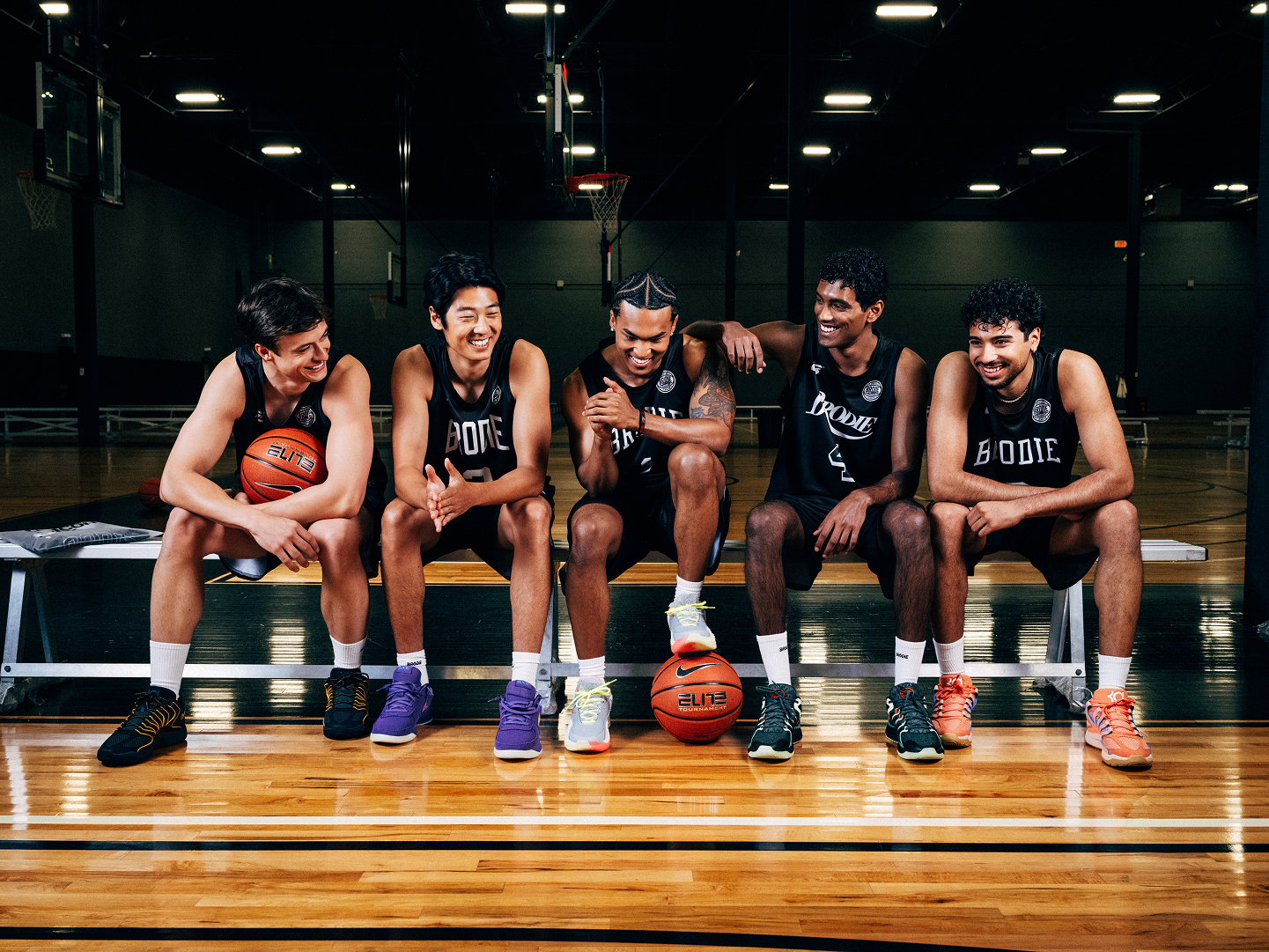 Five basketball players in black Brodie jerseys holding basketballs on an indoor court.
