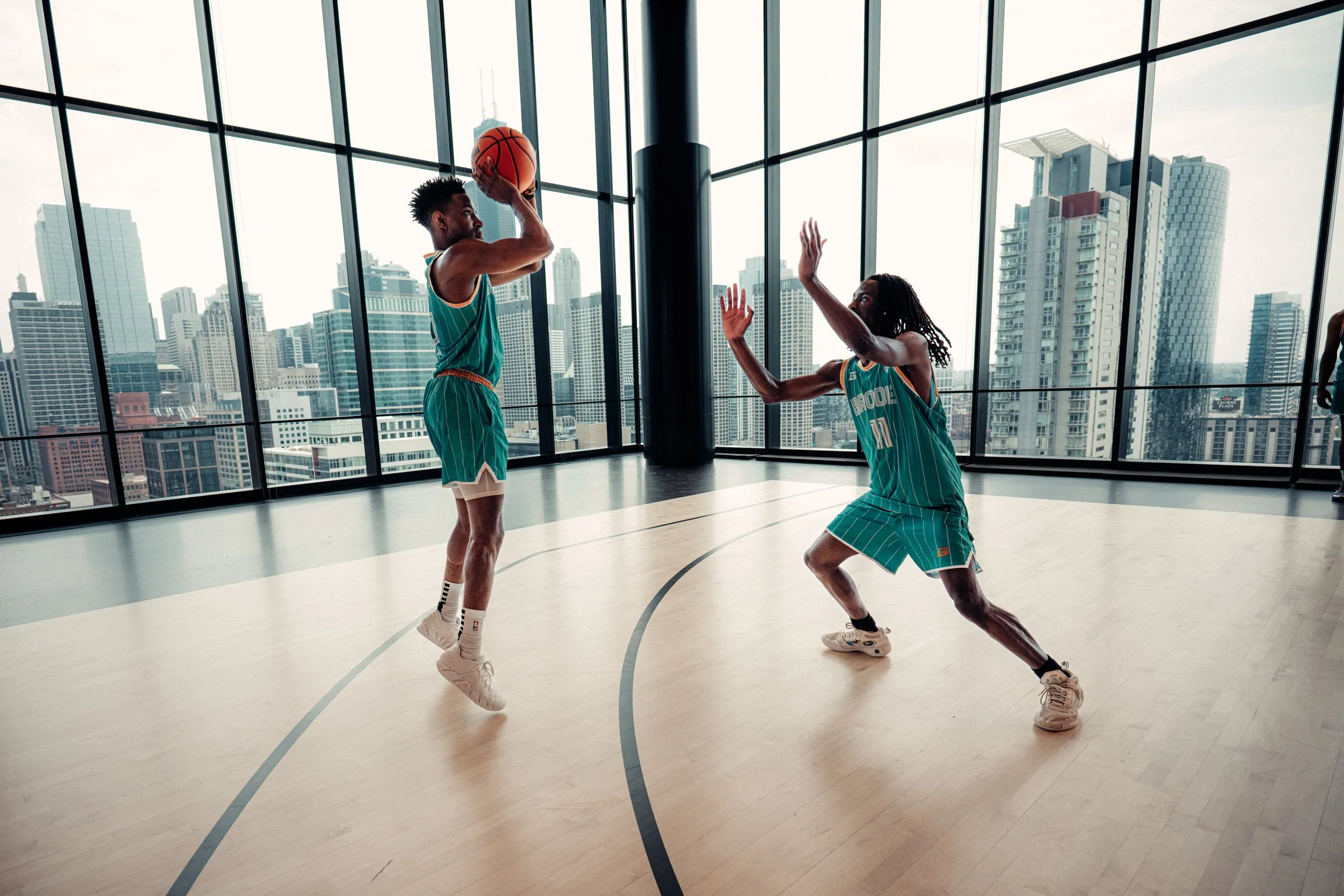 BRODIE athlete in a teal jersey lining up a jump shot in a glass-walled gym above the city