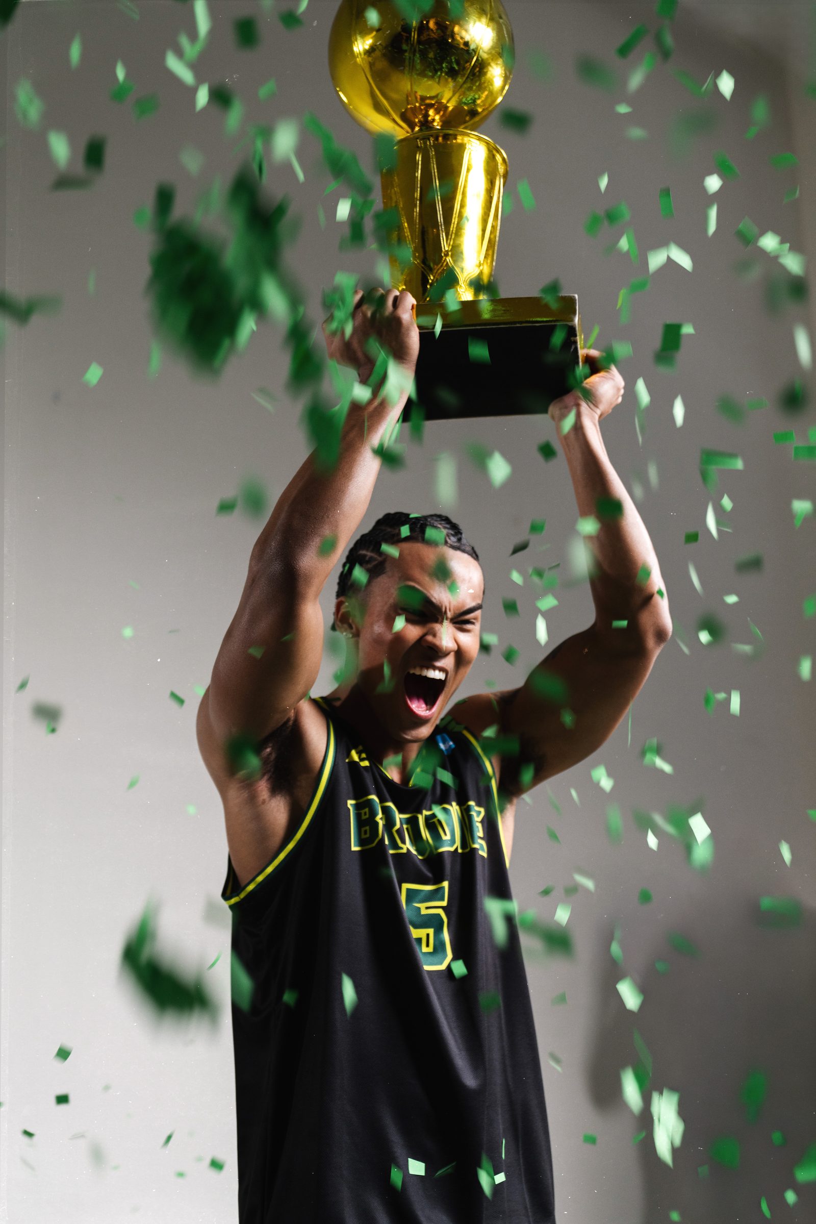 Player in a Brodie jersey roaring with a gold trophy raised overhead as green confetti falls