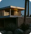 Modern desert-style house at dusk with warm exterior lighting, agave plants, and rocks in the foreground.