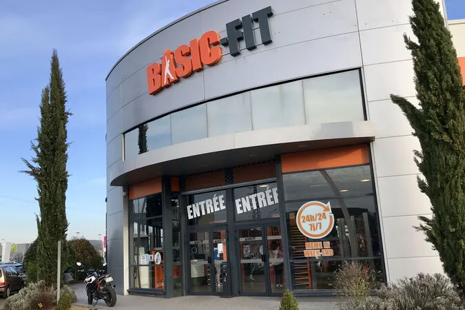 Exterior view of a Basic-Fit gym with glass entrance doors labeled 'ENTRÉE' and a sign indicating 24/7 access, flanked by tall cypress trees.