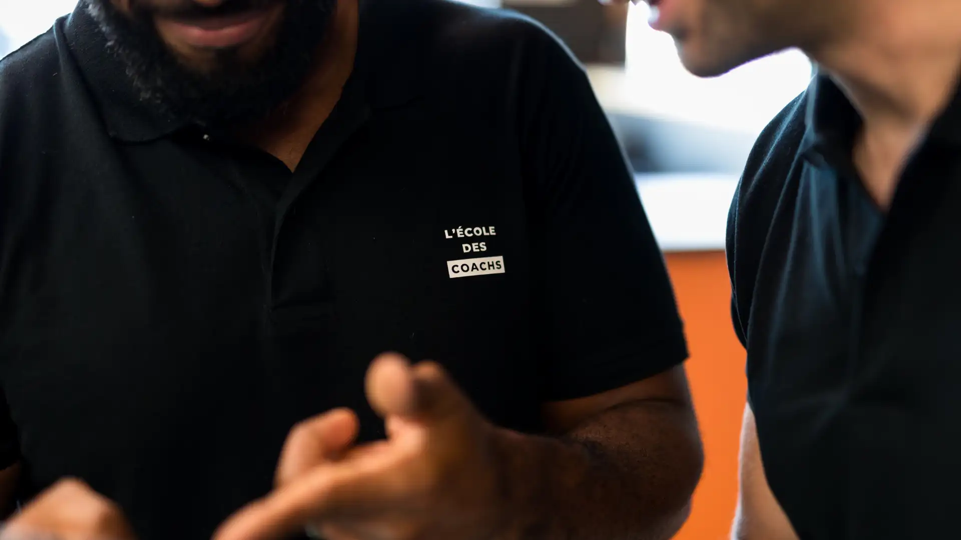 Close-up of two people wearing black polo shirts, one shirt displays the text 'L'ÉCOLE DES COACHS'.