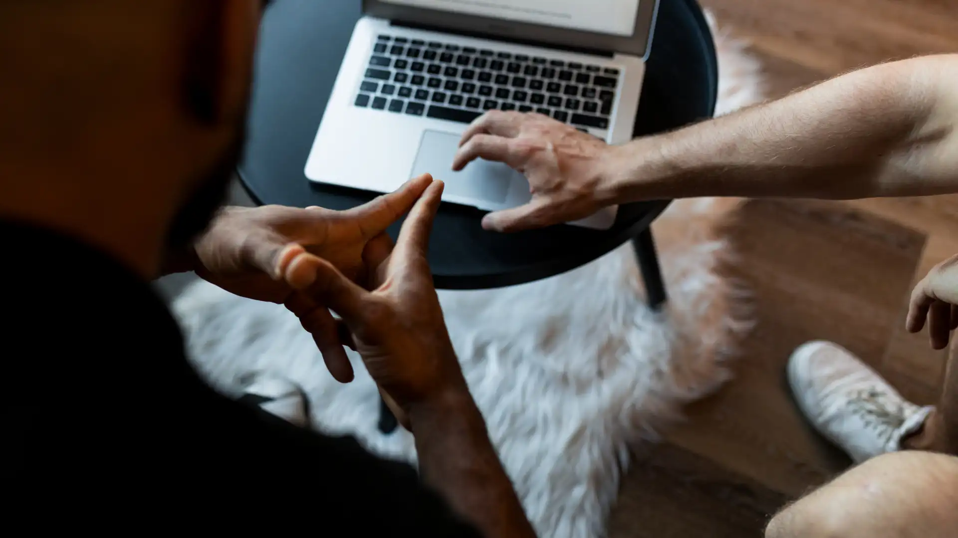 Two people interacting with a laptop on a round black table over a white fur rug on wooden floor.