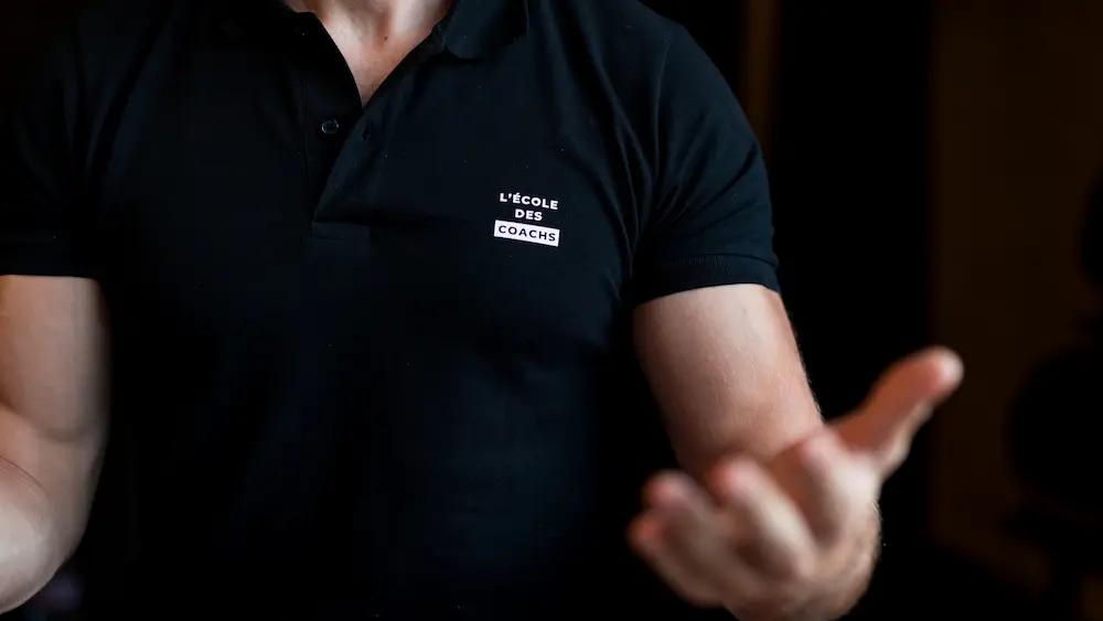 Close-up of a person wearing a black polo shirt with the logo 'L'École des Coaches' on the chest.