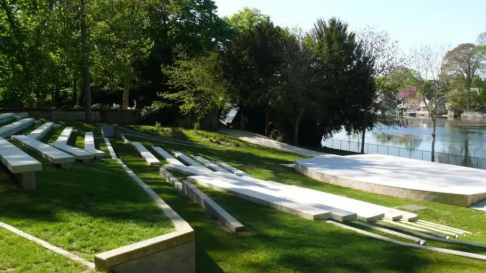 Outdoor amphitheater with white concrete benches on grassy terraced seating next to a river and surrounded by trees.