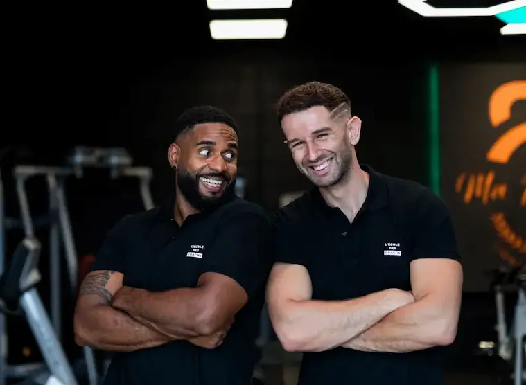 Two male fitness coaches standing with arms crossed, smiling and looking at each other in a gym.