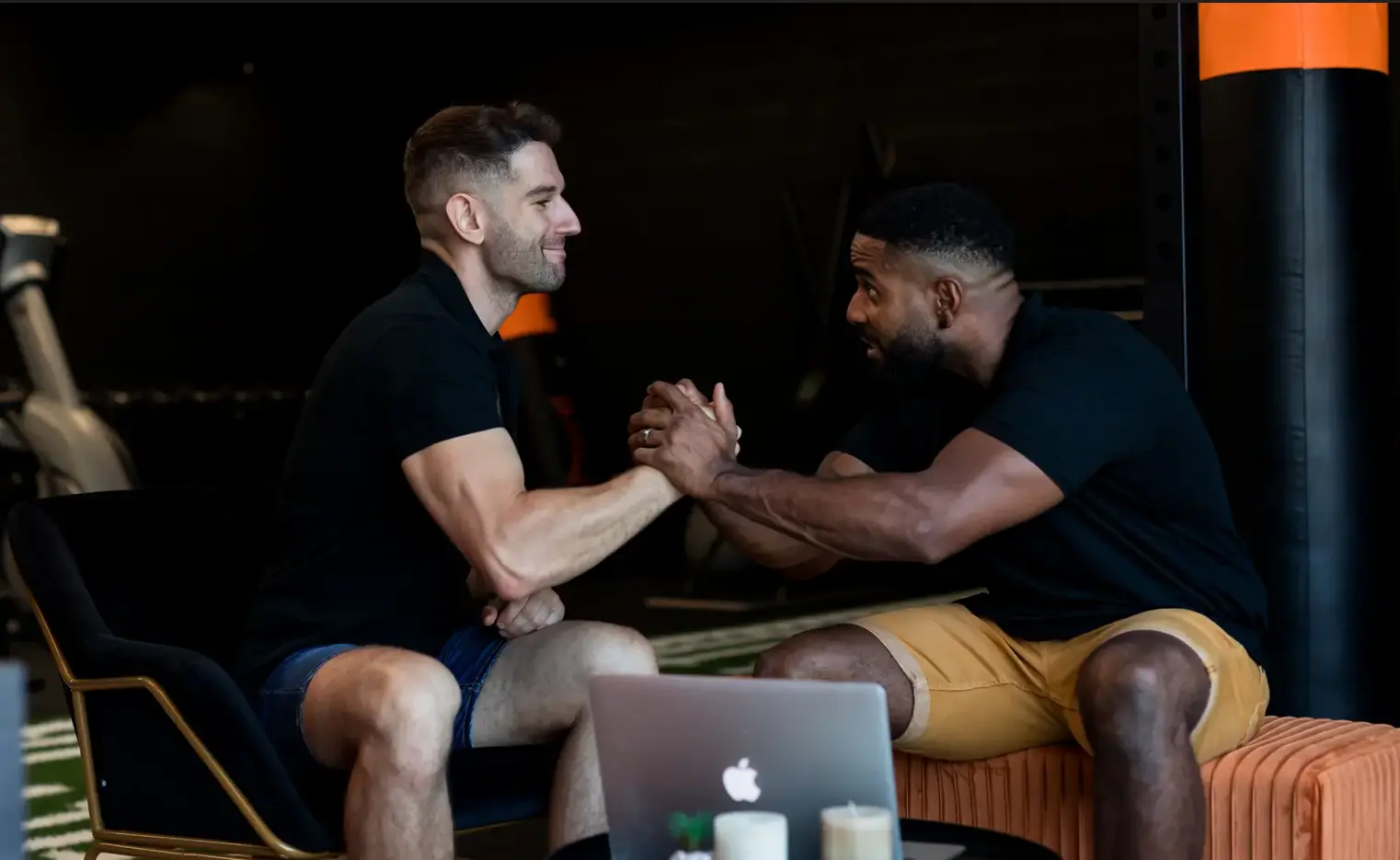 Two men sitting and engaging in a handshake or arm wrestle in a gym setting, with a laptop and candles on a table in front of them.