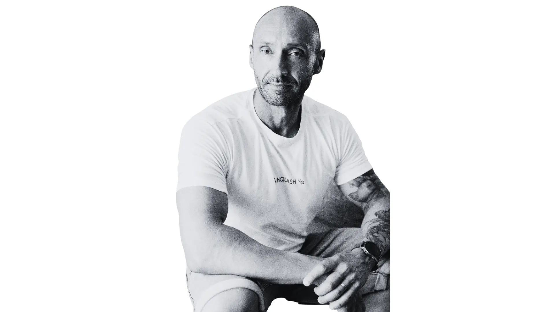 Bald man with tattoos sitting calmly wearing a white t-shirt and shorts against a white background.