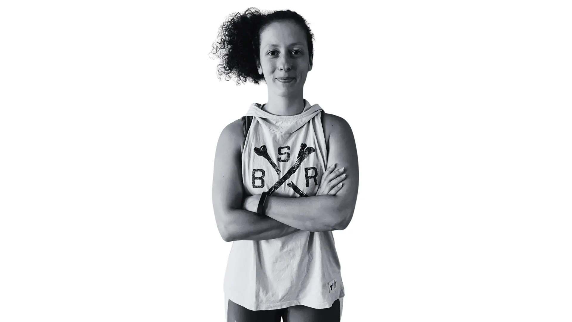 Woman with curly hair tied back, wearing a sleeveless hoodie with crossed bones design and letters B, S, R, standing with arms crossed and smiling slightly.