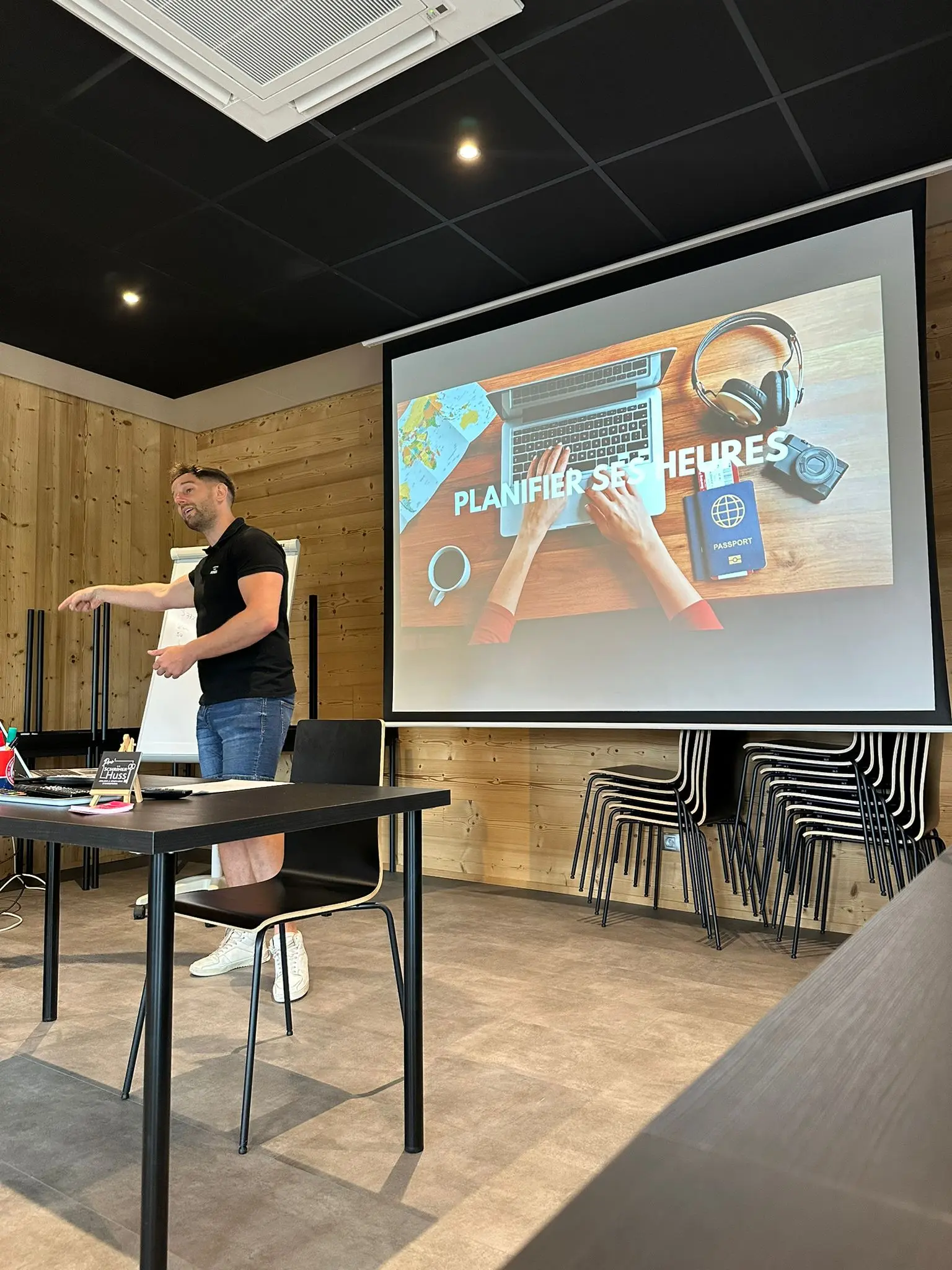 Man in black shirt and denim shorts giving a presentation in a wooden room with a projector screen showing a laptop, passport, headphones, coffee cup, and the text 'Planifier ses heures'.