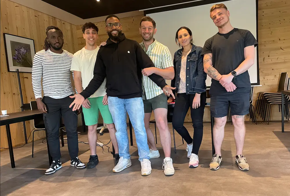 Group of six smiling young adults standing indoors in a casual setting with wooden walls and stacked chairs in the background.