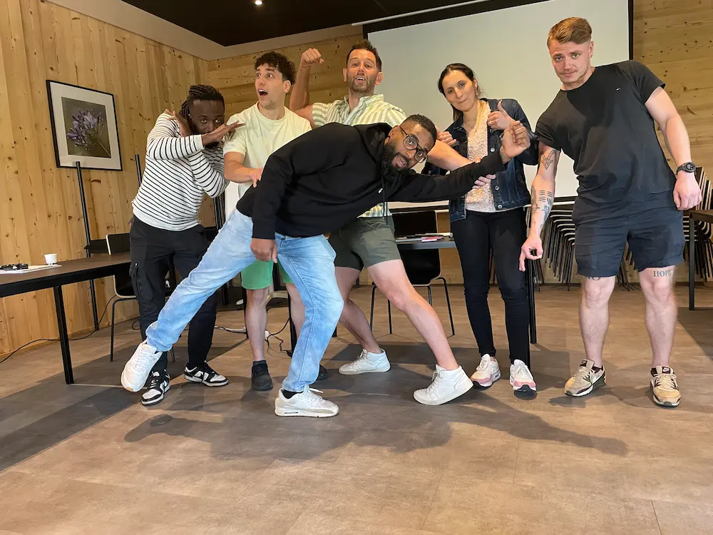 A group of six people indoors striking playful and dynamic poses in front of a projection screen.