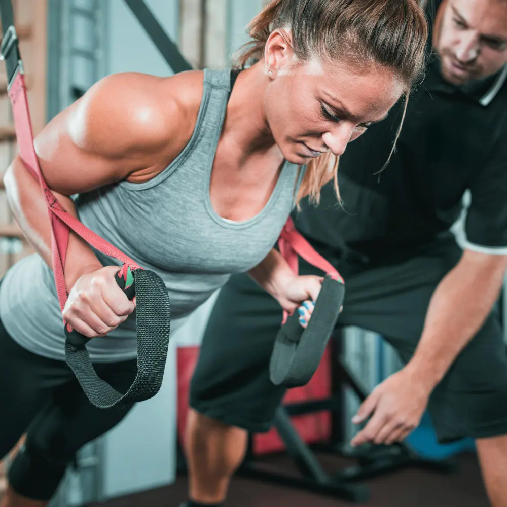 Woman in gray tank top doing suspension training push-ups with a trainer guiding her.