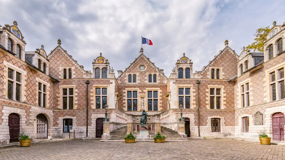 Historic brick building with steep gabled roofs, a French flag on top, and a central statue in the courtyard.