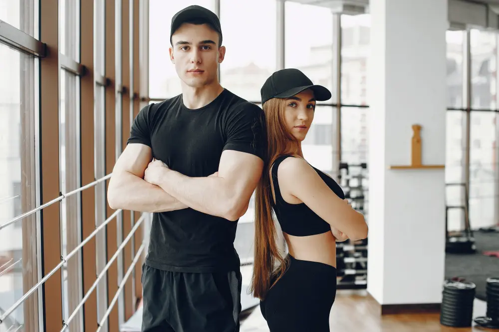 Fit young man and woman wearing black workout clothes and caps standing back-to-back with arms crossed inside a gym.