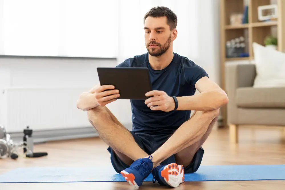 Man sitting cross-legged on a yoga mat indoors, looking at a tablet.