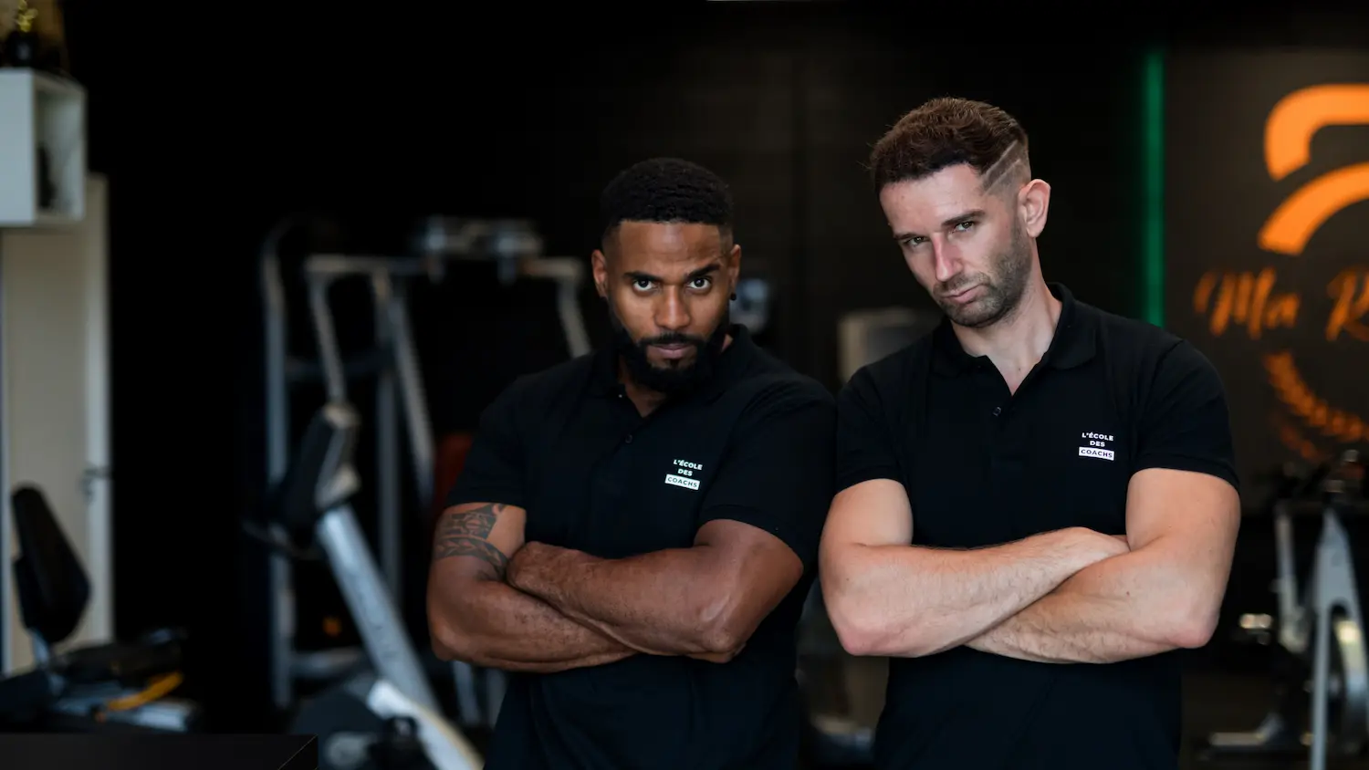 Two men with crossed arms wearing black polo shirts with 'L'ÉCOLE DES COACHS' logo posing confidently inside a gym.