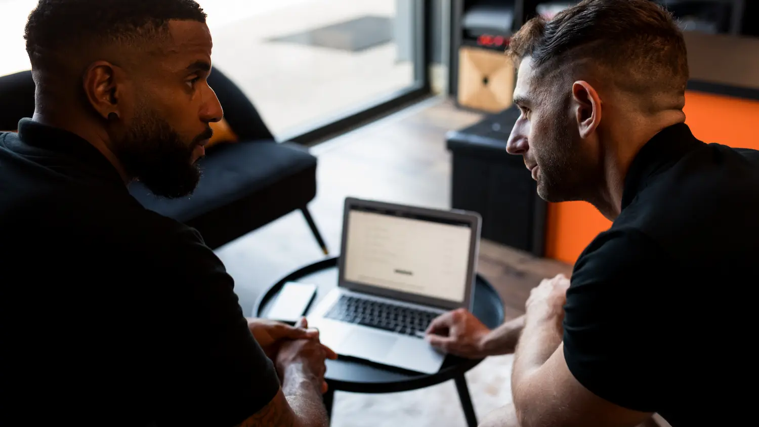Two men sitting at a small round table looking at a laptop screen together indoors.