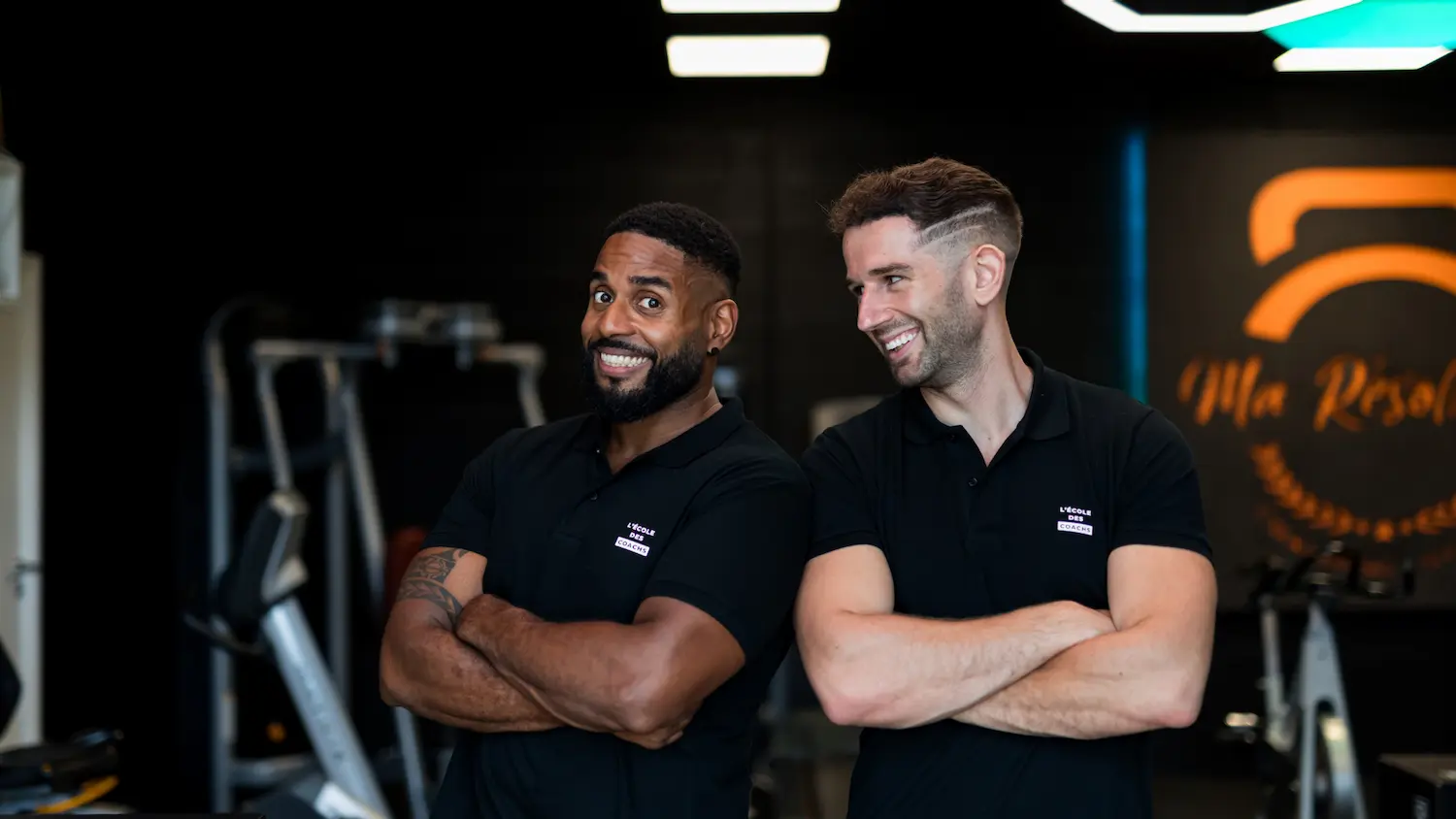 Two male fitness coaches with arms crossed smiling in a gym setting.