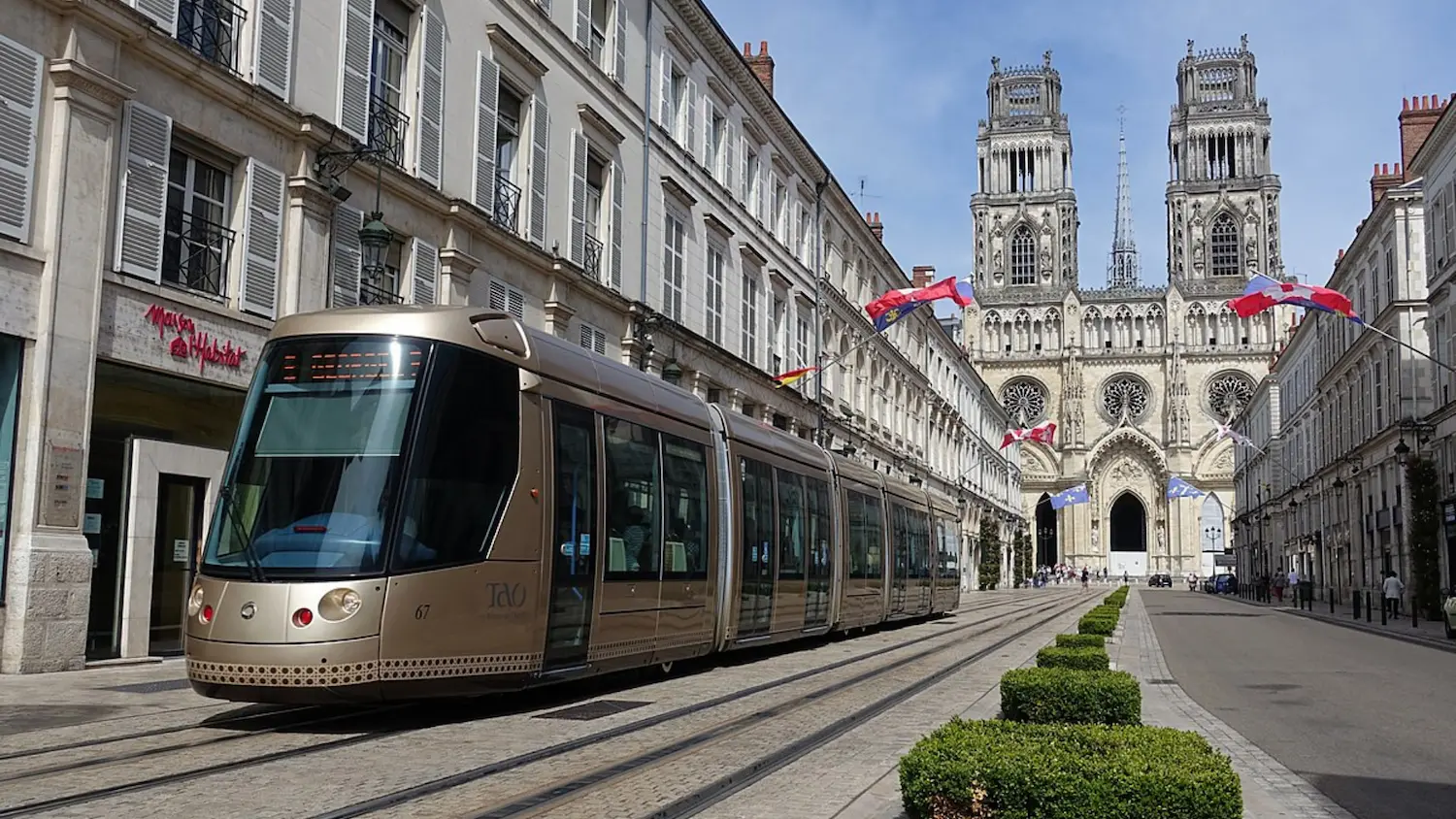 Modern gold tram on stone-paved tracks in a European city street with historic cathedral and flags in the background.