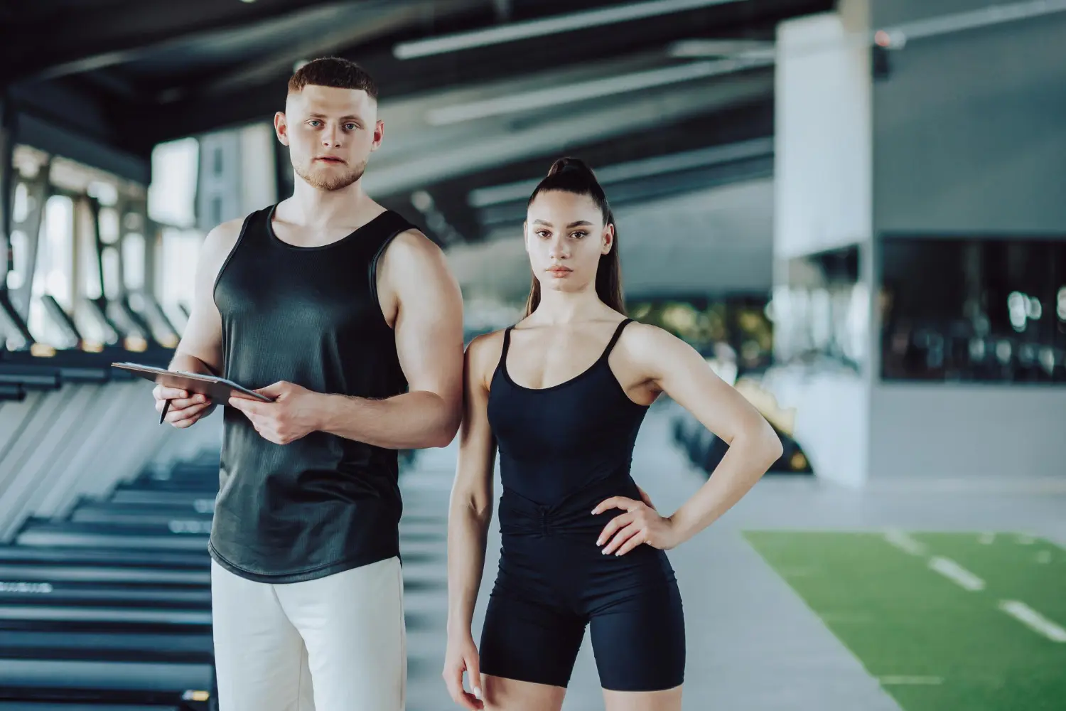 A male fitness trainer holding a clipboard stands beside a female athlete posing confidently in a gym with treadmills in the background.