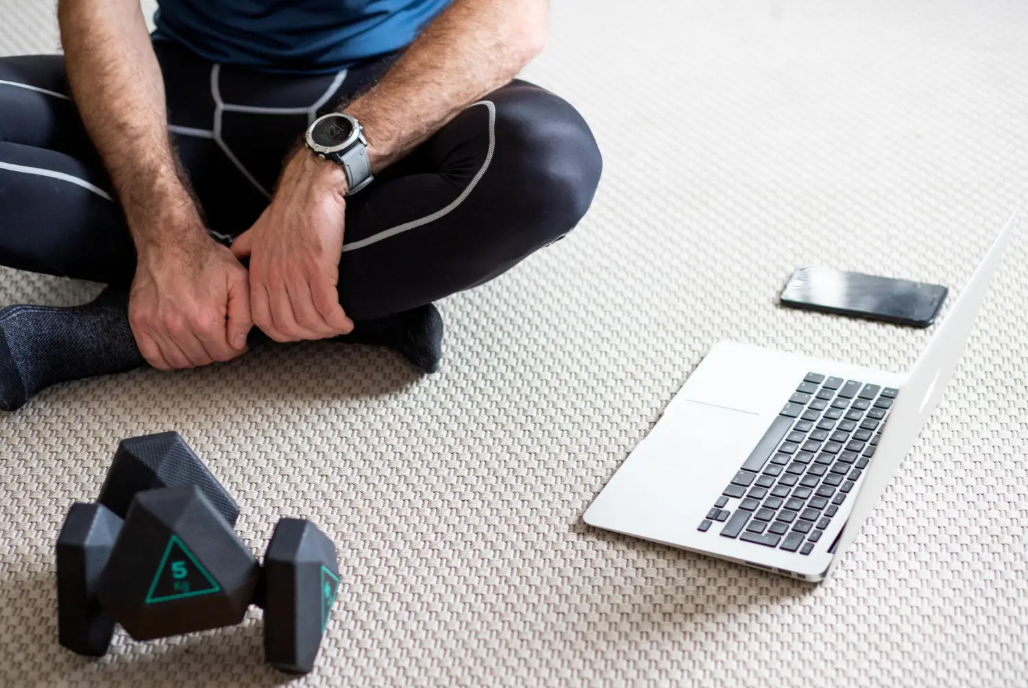 Person in workout clothes sitting cross-legged on a carpet with dumbbells, a smartphone, and an open laptop nearby.